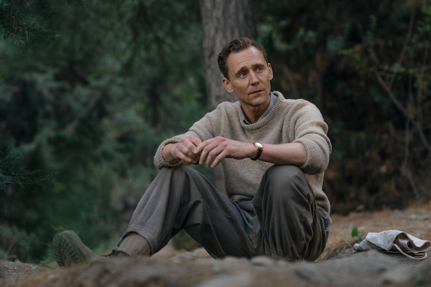 Actor Tom Hiddleston, wearing a beige jumper and grey trousers, sits in a forest gazing into the distance.