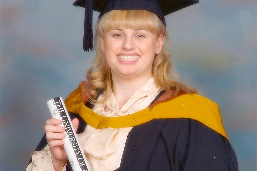 actor rebel wilson wearing a graduation hat, robes and holding a degree smiling at the camera