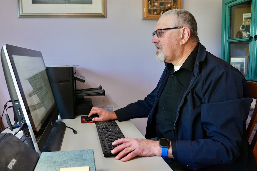 A man with short grey hair and glasses sits at a computer looking confused.