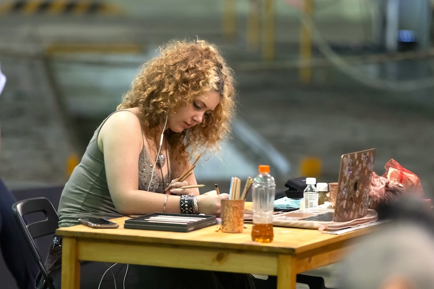A young woman with curly hair stares down at a crowded table with multiple pencils in her hand as she draws, one headphone in