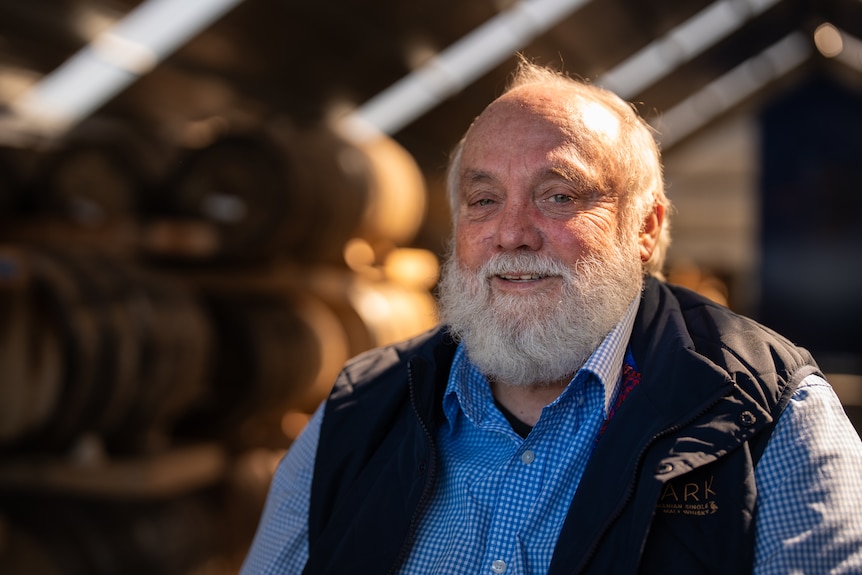 An older man in a blue vest sits in front of whisky barrels in a large shed