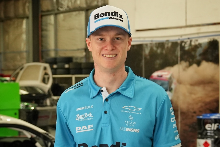A young man wearing a supercar racing jersey and cap stands smiling in front of a V8 supercar in a workshop.
