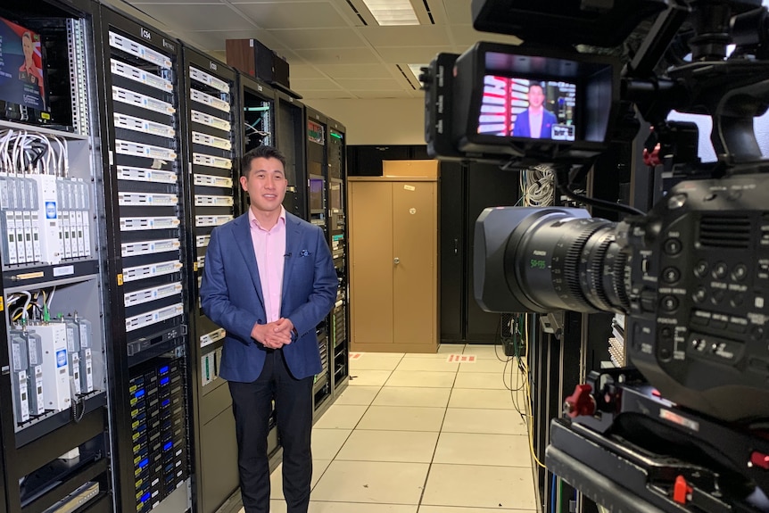 David standing in a server room, a few metres away from a video camera on a tripod filming him.