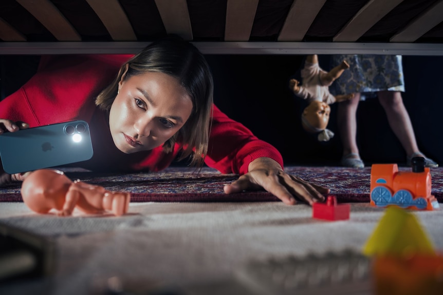 Tuuli Narkle, an Aboriginal woman in her late 20s, is holding an iPhone with its light on up as she looks under a bed.
