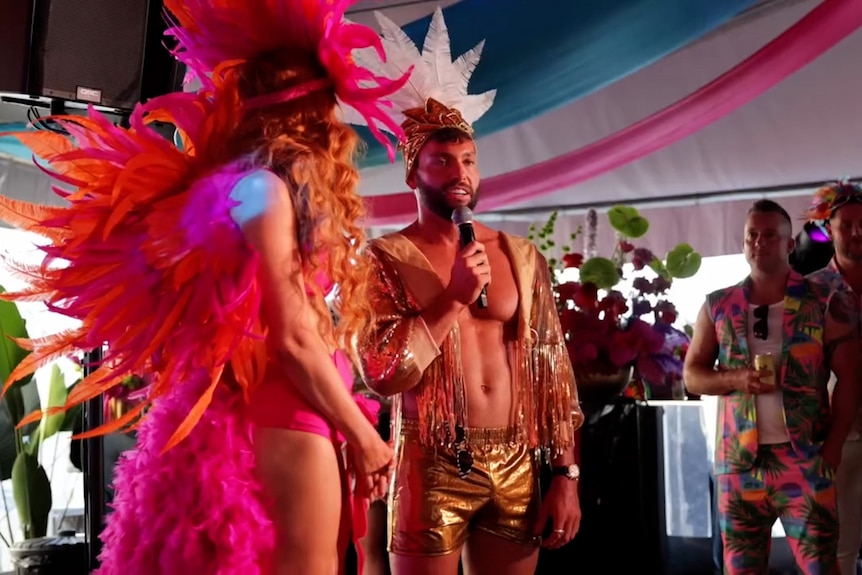 a man wearing gold shorts and a decorative headpiece speaks into a microphone as a carnival dancer and party guests look on 