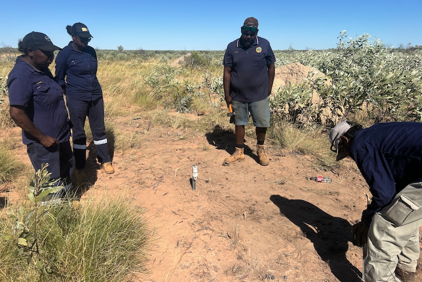 Four rangers dressed in navy look at a camera set up on red dirt in desert.