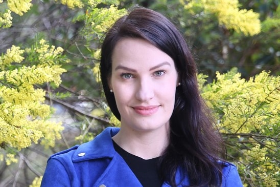 A woman with long dark hair and her arms crossed, wearing a vibrant blue jacket, looks straight at the camera.