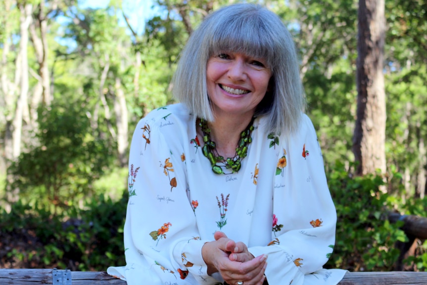 Bernice Barry, an older woman, with a grey bob cut with a fringe, sits outside, hands clasped, head tilted.