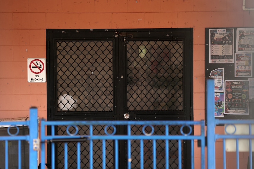Double doors, with security screens on them, on a brick building. A pinboard is on the wall. A low fence is in the foreground.