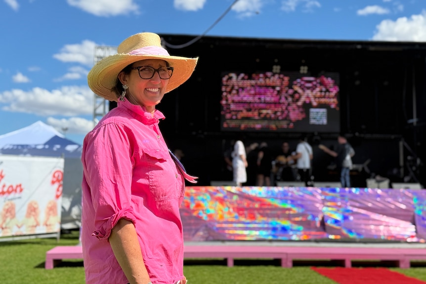 A woman in a broad-brimmed straw hat wearing a pink collared shirt stands in front of a stage smiling on a sunny day.