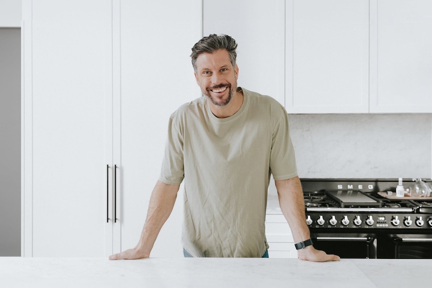 Man (Sam Wood) in beige T-shirt leaning on a kitchen bench with wide smile.