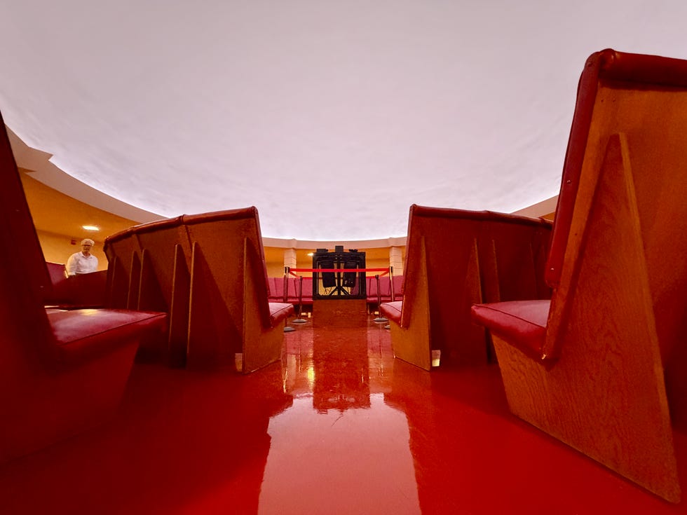 Interior view of a circular room featuring red benches and a circular ceiling, with a person standing in the background.