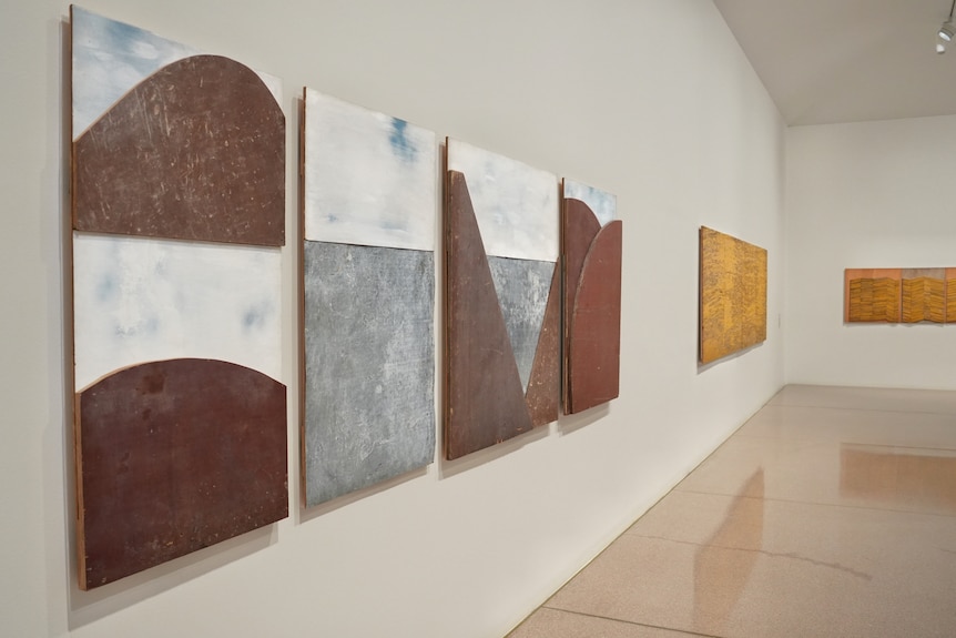 Four artworks on a wall as the focus that are brown and grey, two yellow artworks in the background.