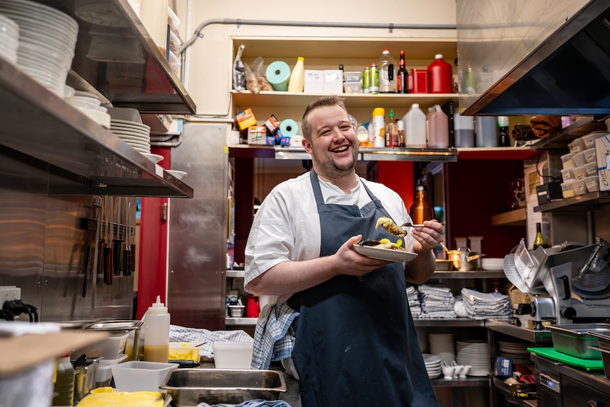 A man in a white shirt and blue apron stands in a cluttered kitchen holding a plate of food