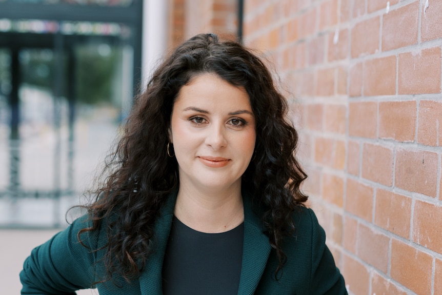 A portrait of a woman with brown curly hair wearing a green blazer standing in front of a brick wall.