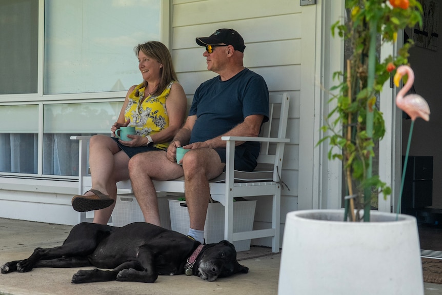 A couple sit on a white bench outside a home as a black dog lounges beside them. 
