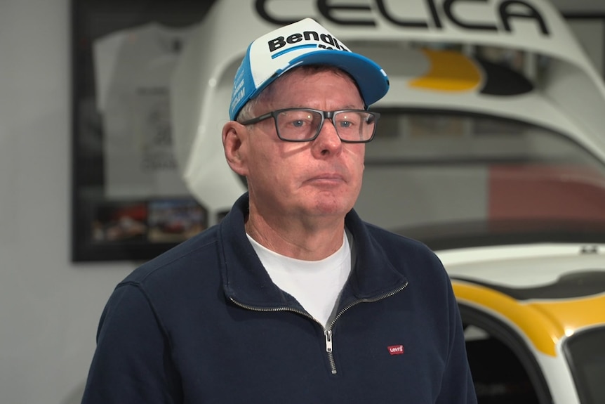 A man wearing a motorsports cap stands in front of a supercar speaking.