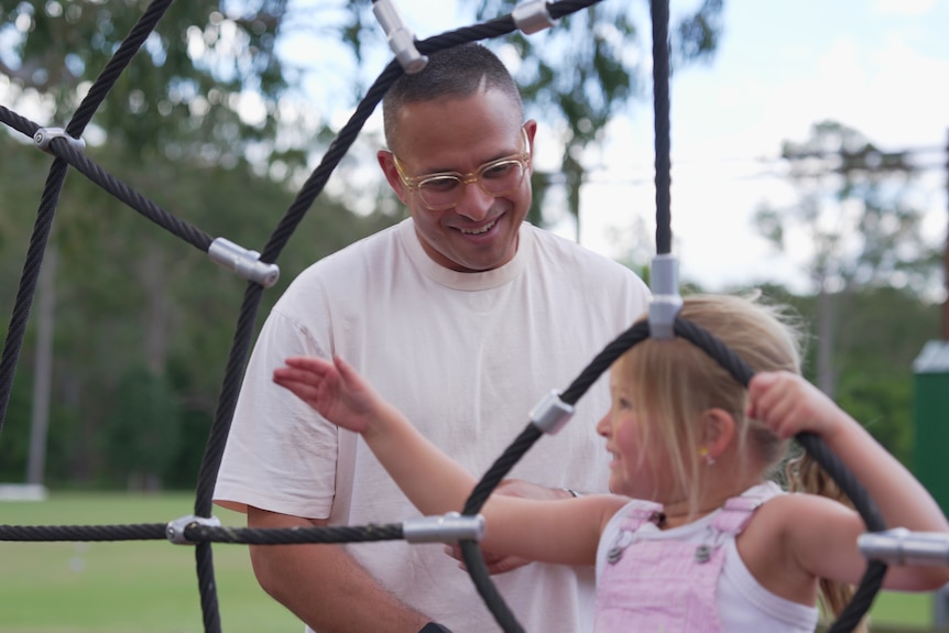 Usman Khawaja, wearing glasses, looks down at Ayla and smiles as she holds onto play equipment.