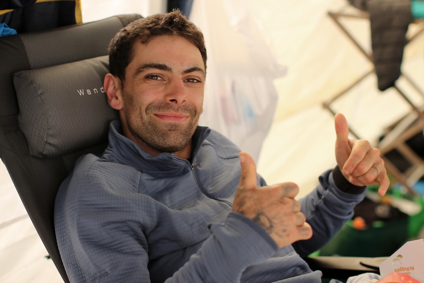 Brandon Lee sits on a chair in a blue jumper giving a thumbs up and smiling.