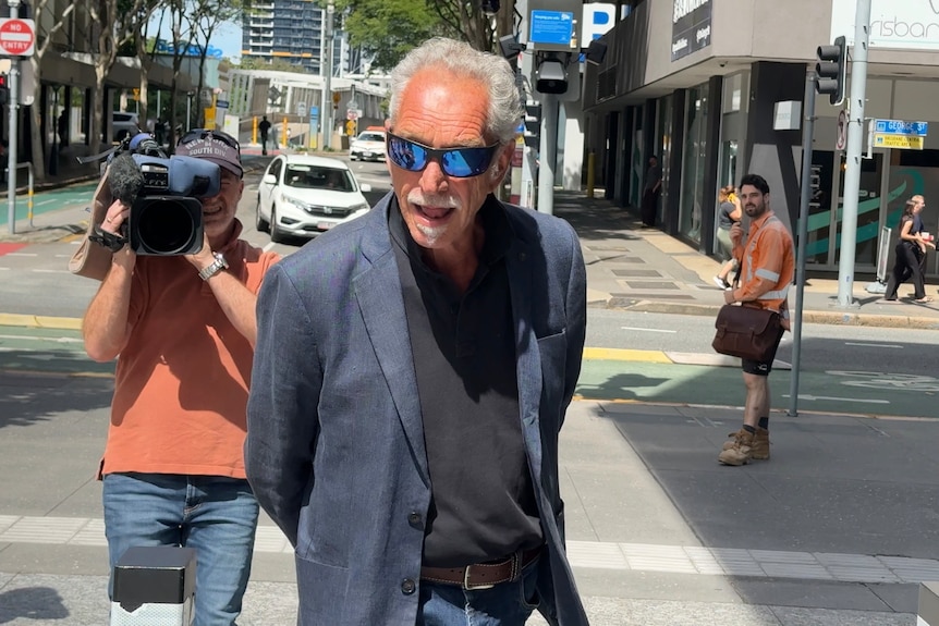 A man in sunglasses wearing a suit walking with a TV camera behind him.