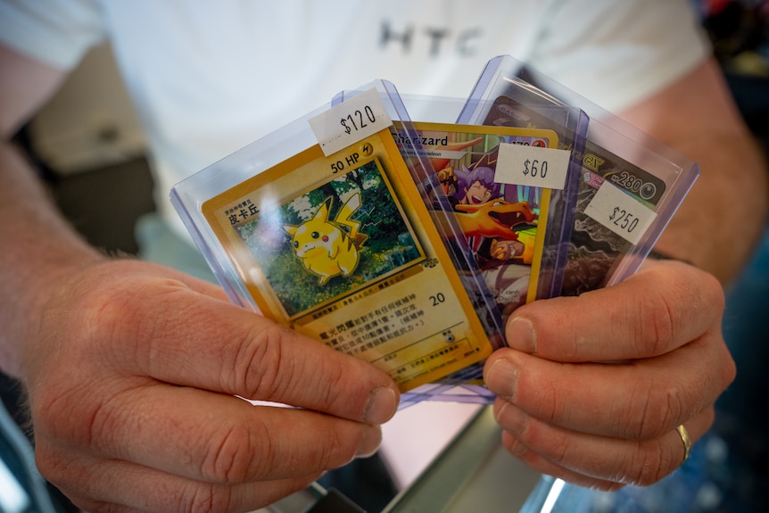 Hands holding Pokemon cards each priced at $120, $65 and $250