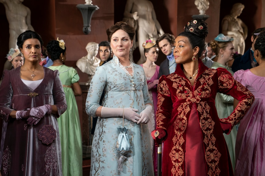 Three women in front of a party of people all wearing colourful period costumes with hair tied back.
