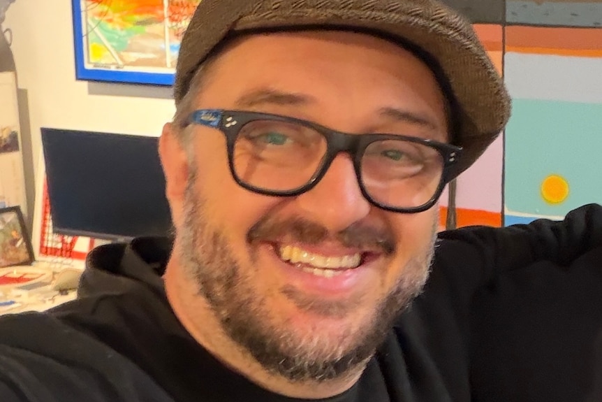 A man wears a glasses and cap and smiles.