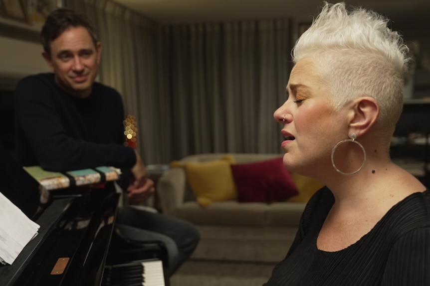 Seated Katie Noonan with short blonde, styled hair seated at a piano, mid-sentence, as Trent Dalton sits nearby listening.