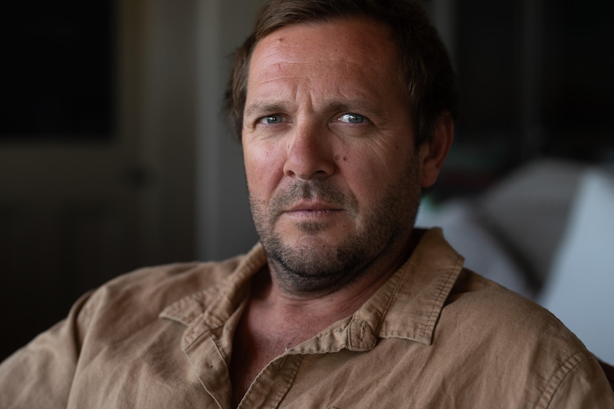 A dark-haired man sits in a softly-lit room.