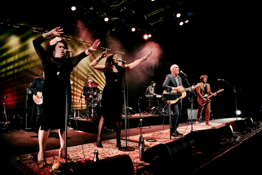 Vika and Linda sing live on stage alongside Paul Kelly and a band guitarist