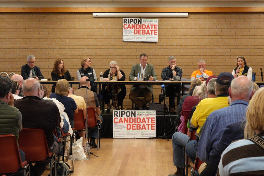 Eight men and woman sit at a long table in front of a crowd in a community hall.