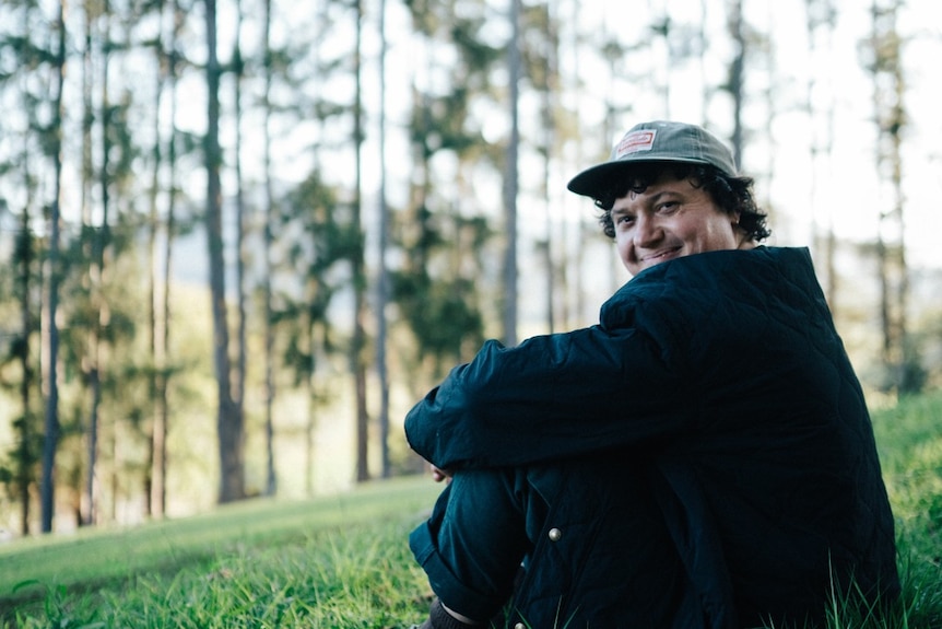 A man sits on the grass looking over his shoulder in front of trees