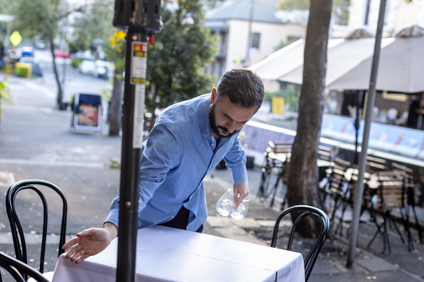 A man in a blue shirt sweeps crumbs off a white tablecloth on a restaurant table set up on the footpath