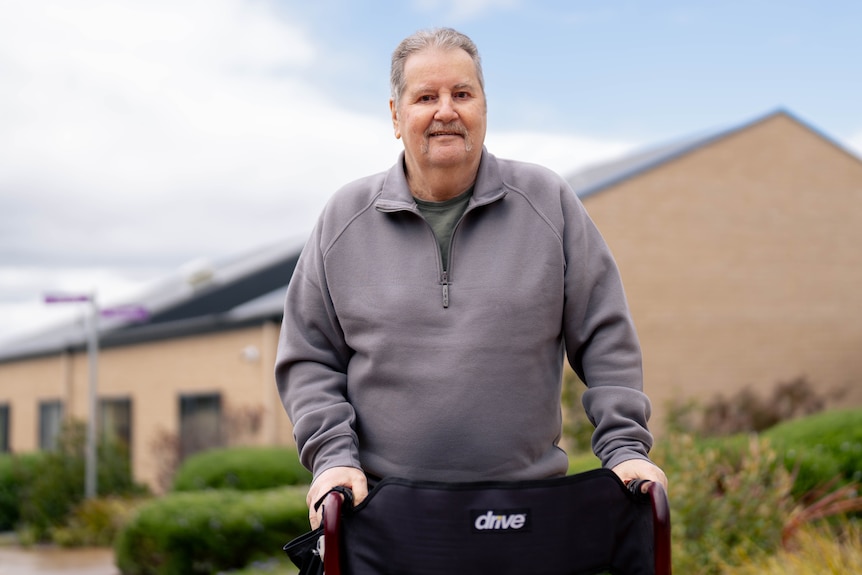 Nick, wearing a grey quarter zip jacket, stands with a walker smiling for a photo with aged care housing behind him.