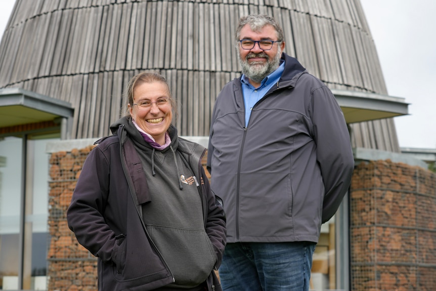 Evelyn Nicholson and Damein Bell stand smiling in front of a stone hut.