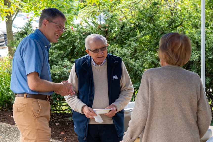 A man in a blue shirt and tan pants smiling while talking with an older man and woman in an outdoor picnic area.