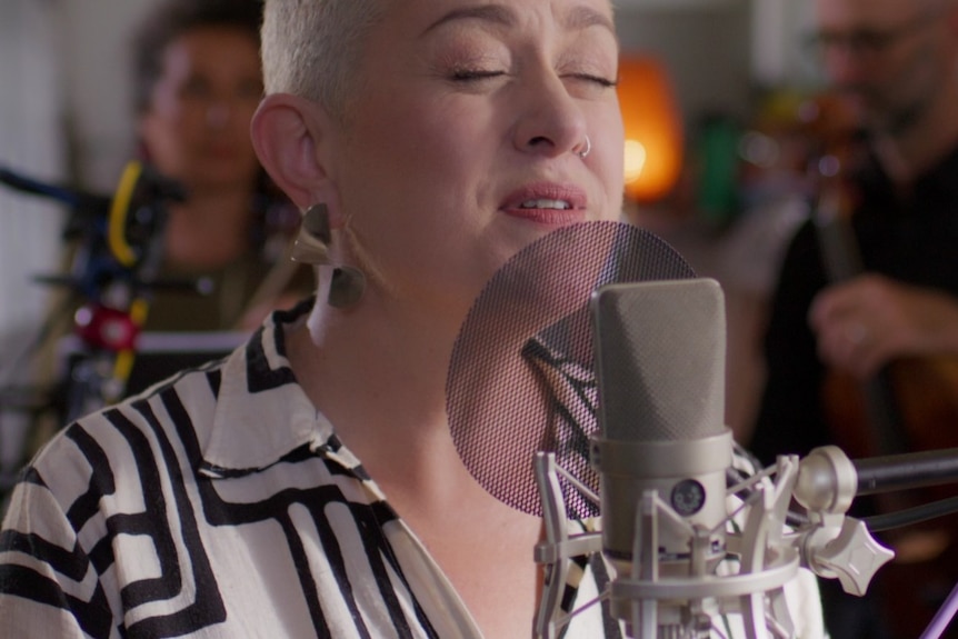 Katie Noonan with short blonde hair wears black and white top mid-singing into a microphone.