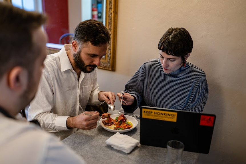 Two people sit at a table and taste a plate of food with a chef watching