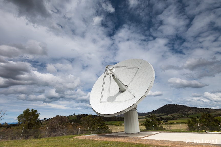 Large white telescope antenna against a green hilly and cloudy backdrop.
