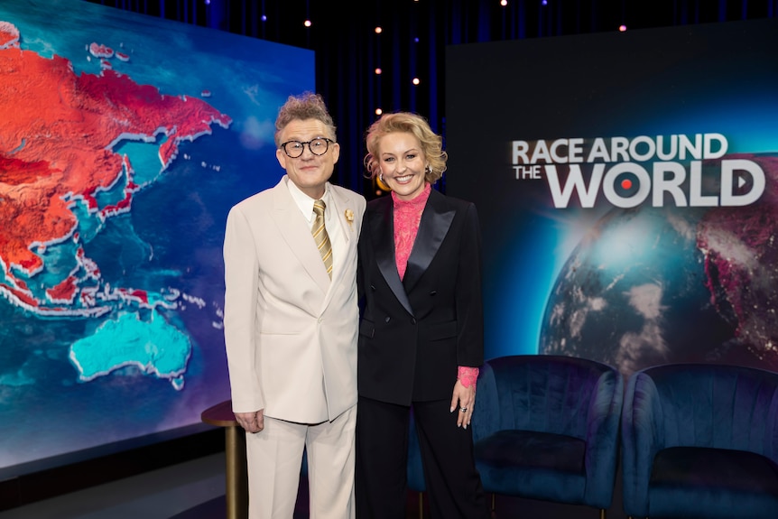 John and Zan beam as they stand in front of a studio background depicting a world map and the logo for their show.