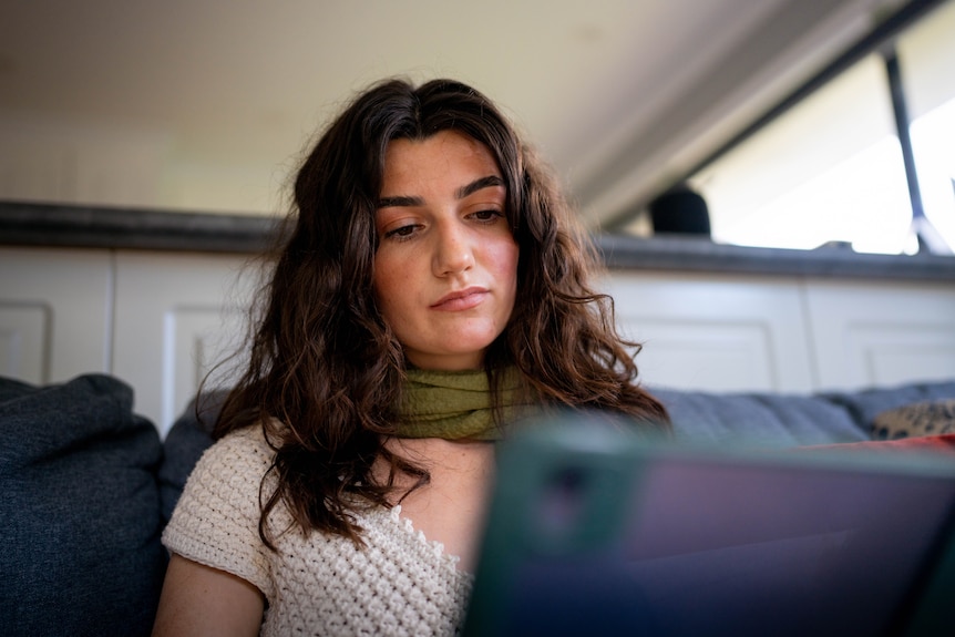 Lucy, wearing a crochet top and a green scarf, sitting on a couch holding an iPad with a serious expression.