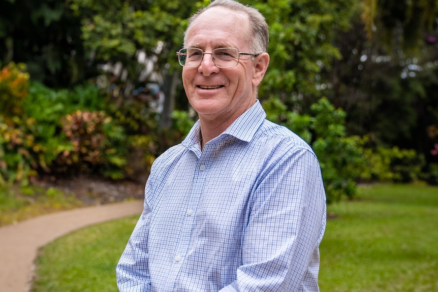 An older gentleman with glasses stands proud in front of a lush garden.