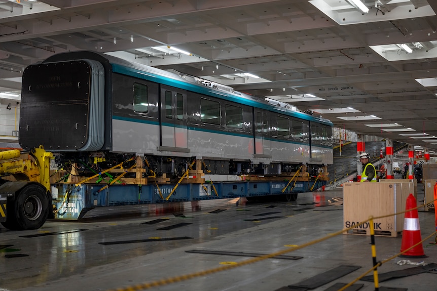 A three-carriage-long train with black, silver and teal detailing loaded onto a long trailer on a cloudy day.