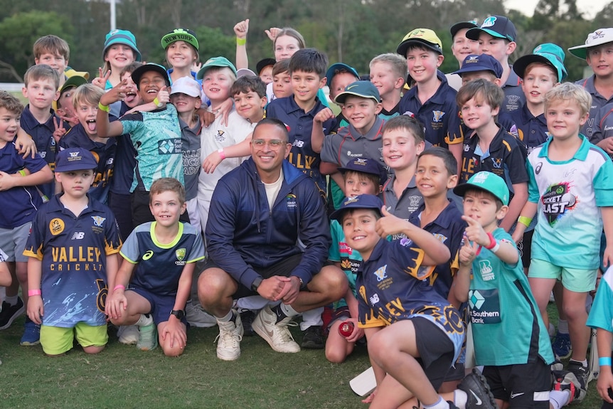 Cricketer Usman Khawaja kneels down in front of a large group of excited children.