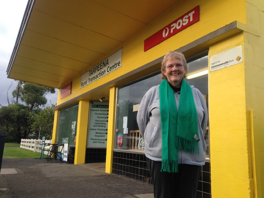 A mature woman wearing a green scarf and with her hands behind her back stands outside a yellow post office.