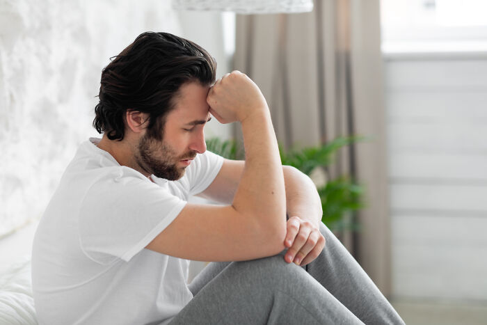 Anxious man sitting on a bed resting his head on his hand while feeling sick.