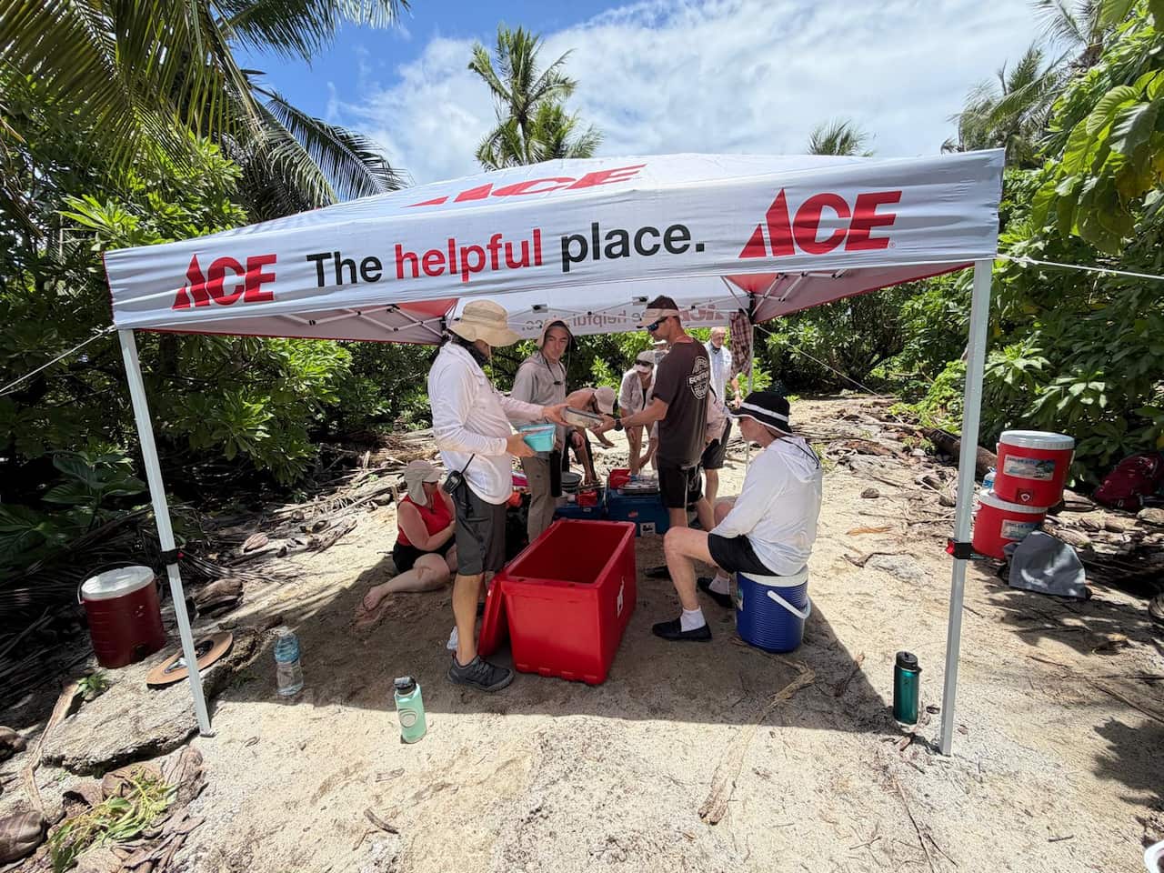 Researchers shelter beneath a white ACE Hardware canopy on a sandy beach surrounded by dense tropical vegetation on Nikumaroro Island. Several people sit or stand around red storage tubs and coolers while sorting equipment in the shade, with palm trees and blue sky visible overhead.