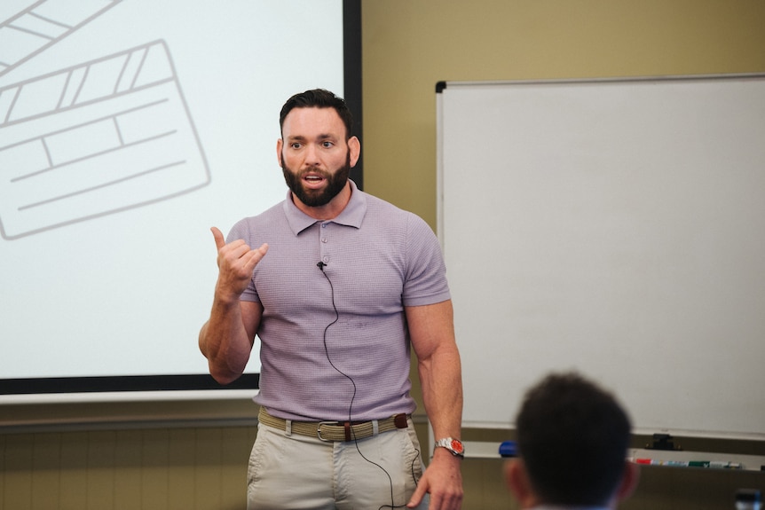 a fit man wearing a tight purple collared shirt and chinos talking in front of a whiteboard