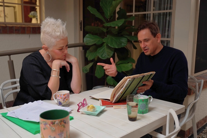 Katie Noonan leans over her hand at a table looking down at a notebook that Trent Dalton is flicking through the pages.