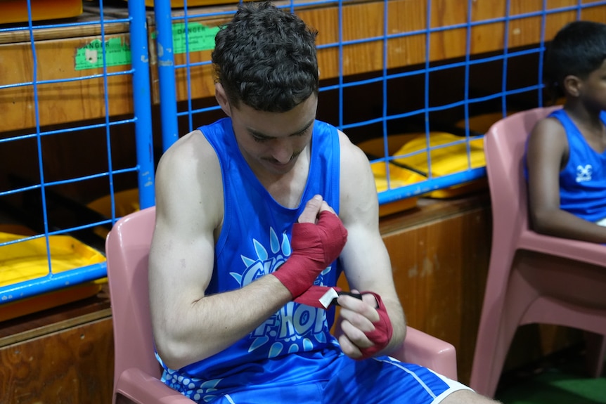 Declan Unwin-Walker wears a blue Gloves of Hope uniform inside a stadium, he wraps a strip of cloth around his hands and wrist.